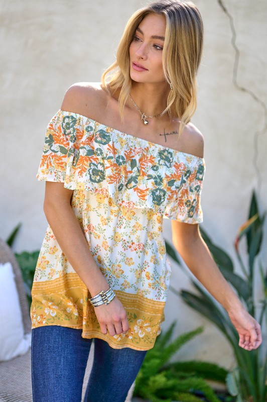 A woman with blonde hair is smiling while standing outdoors, wearing the Printed Off Shoulder Smocked Top paired with jeans.