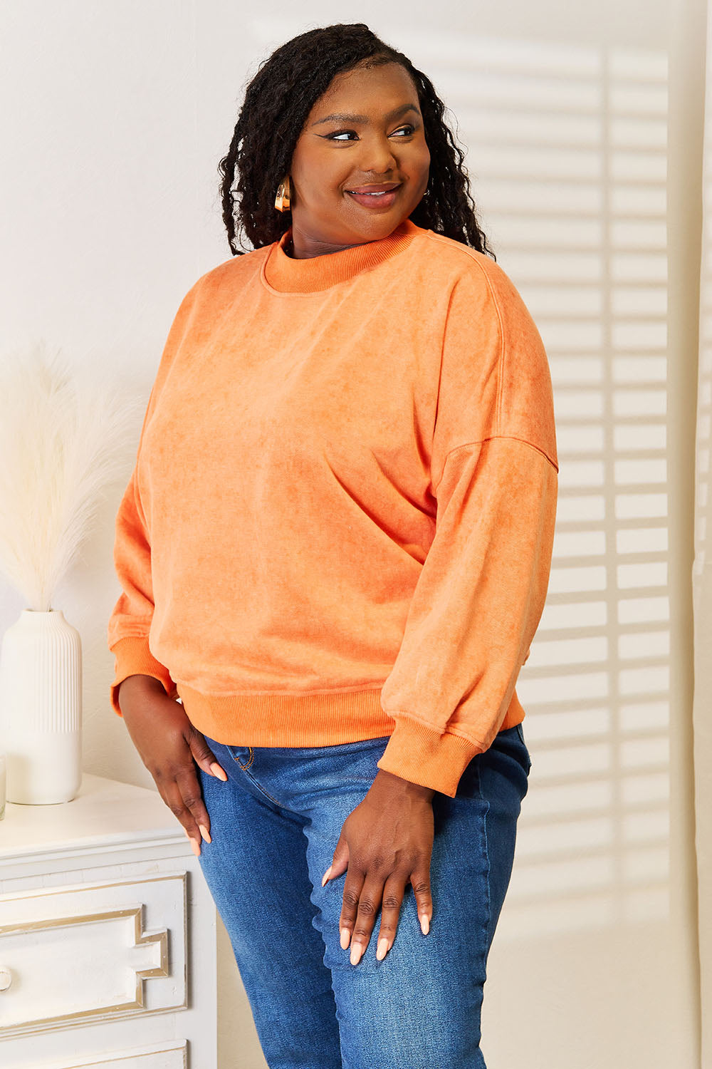 A woman with long, wavy hair smiles in a casual wardrobe, sporting the Basic Bae Round Neck Dropped Shoulder Sweatshirt in orange and blue jeans. She stands beside a white dresser adorned with decorative vases.