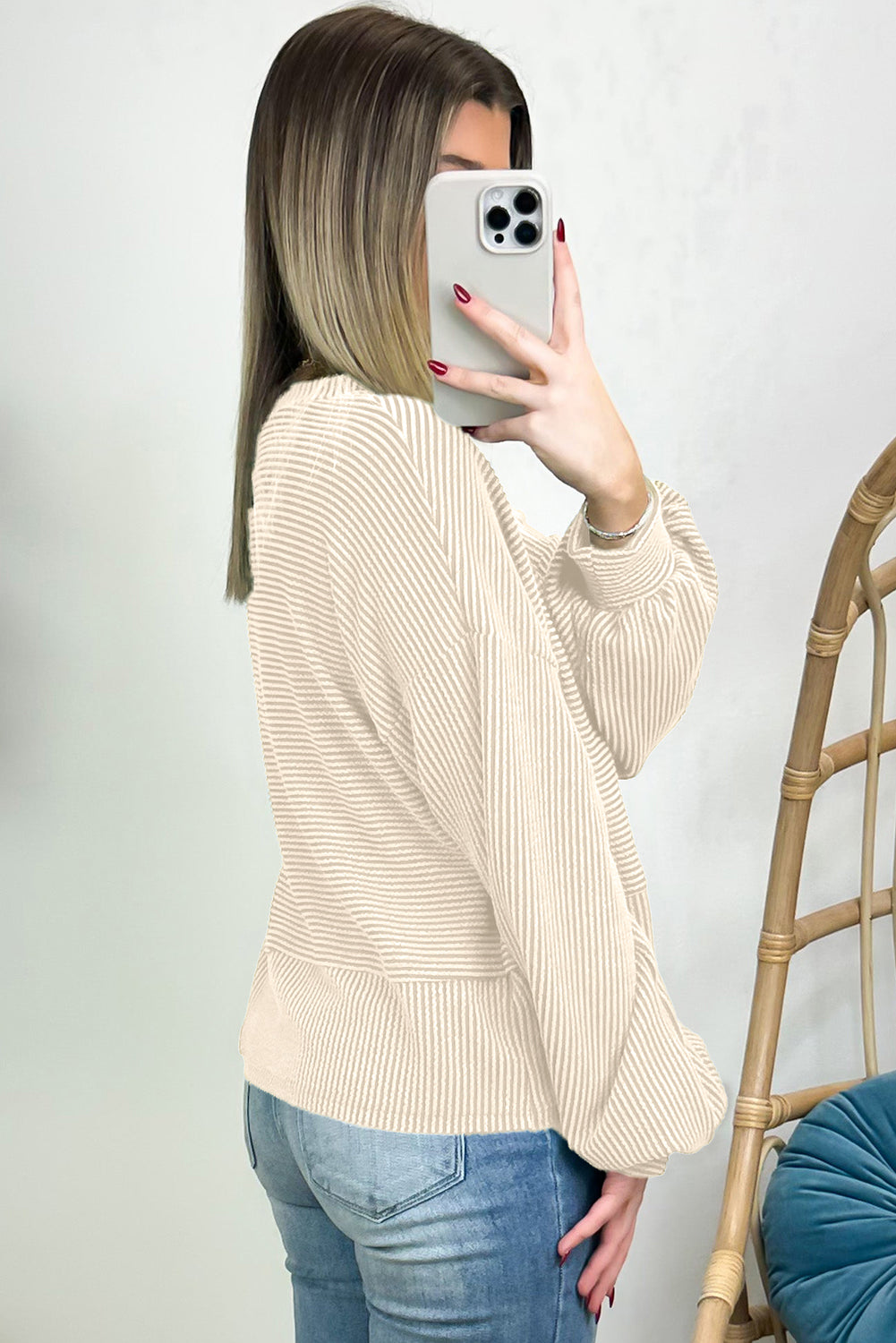 Person wearing the Apricot Corded Texture Lantern Sleeve Buttons Henley Sweatsuit and blue jeans, taking a mirror selfie with a smartphone. Hair is straight and shoulder-length; they stand in front of a white wall and a wooden chair.