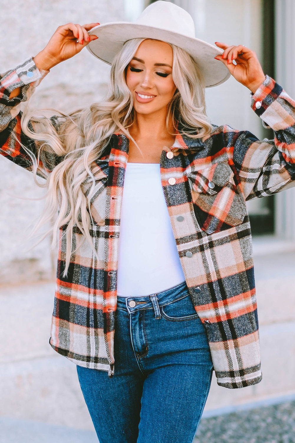 A person wearing a Brown Plaid Button Up Long Sleeve Flannel Shacket layered over a white top with ripped jeans and holding a brown handbag stands against a textured gray wall.