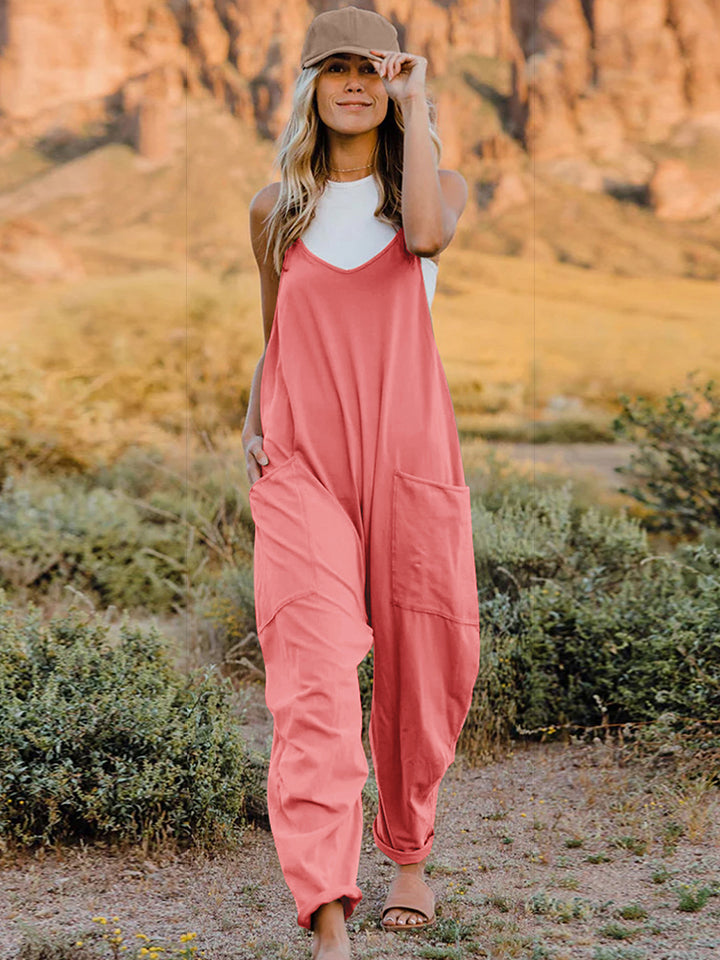 A woman wearing the Double Take Full Size Sleeveless V-Neck Pocketed Jumpsuit in coral and a beige cap is walking outdoors on a clear day, with a rocky landscape in the background.