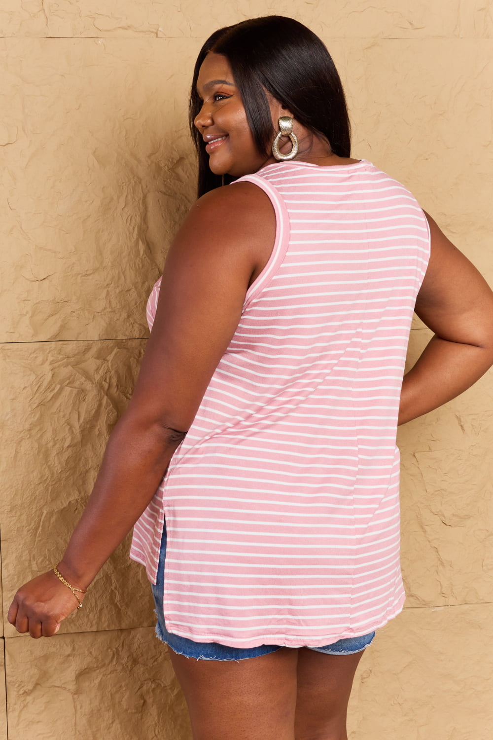 A woman wearing the Doublju Talk To Me Full Size Striped Sleeveless V-Neck Top in pink and white, paired with denim shorts, smiles while standing against a textured beige background.