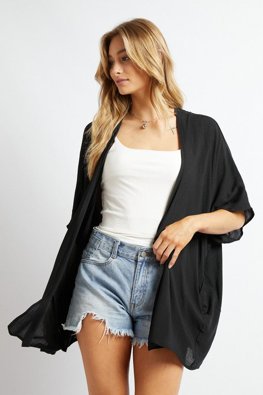 A woman poses against a simple backdrop, gracefully wearing a wardrobe essential: the Favorite Solid Kimono Cardigan in pink. Complementing her white tank top and denim shorts, this casual layer enhances her relaxed style with an elegant touch.