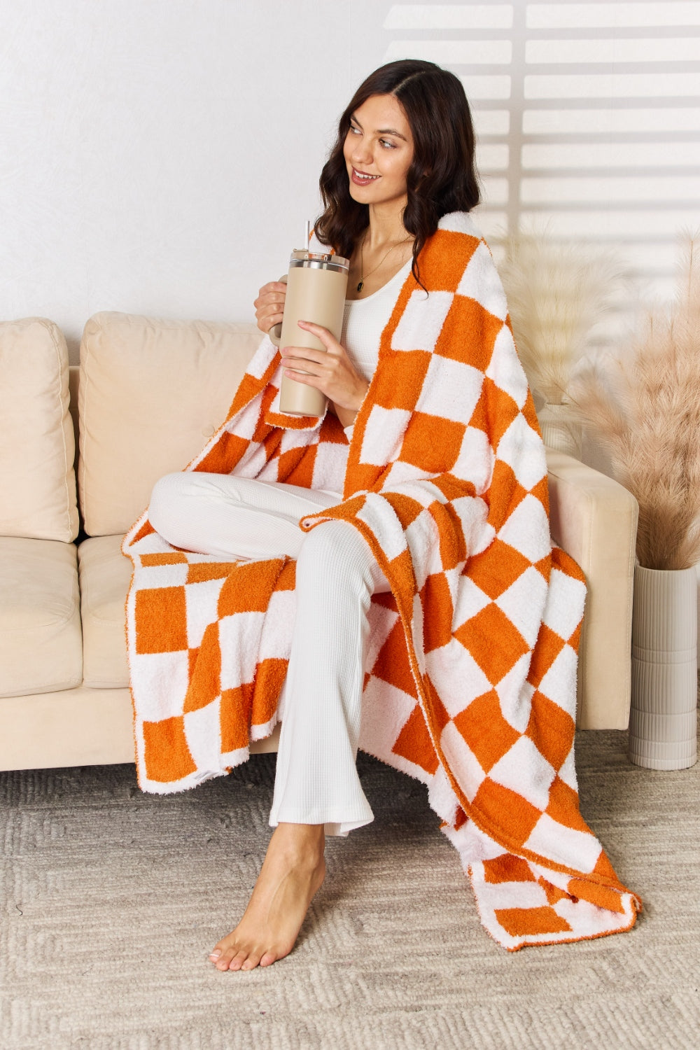 A person stands holding a large Cuddley Checkered Decorative Throw Blanket, smiling, in a room with a sofa and a potted plant. The blanket features a black and white checkerboard pattern, is made from 100% polyester, and imported for quality.