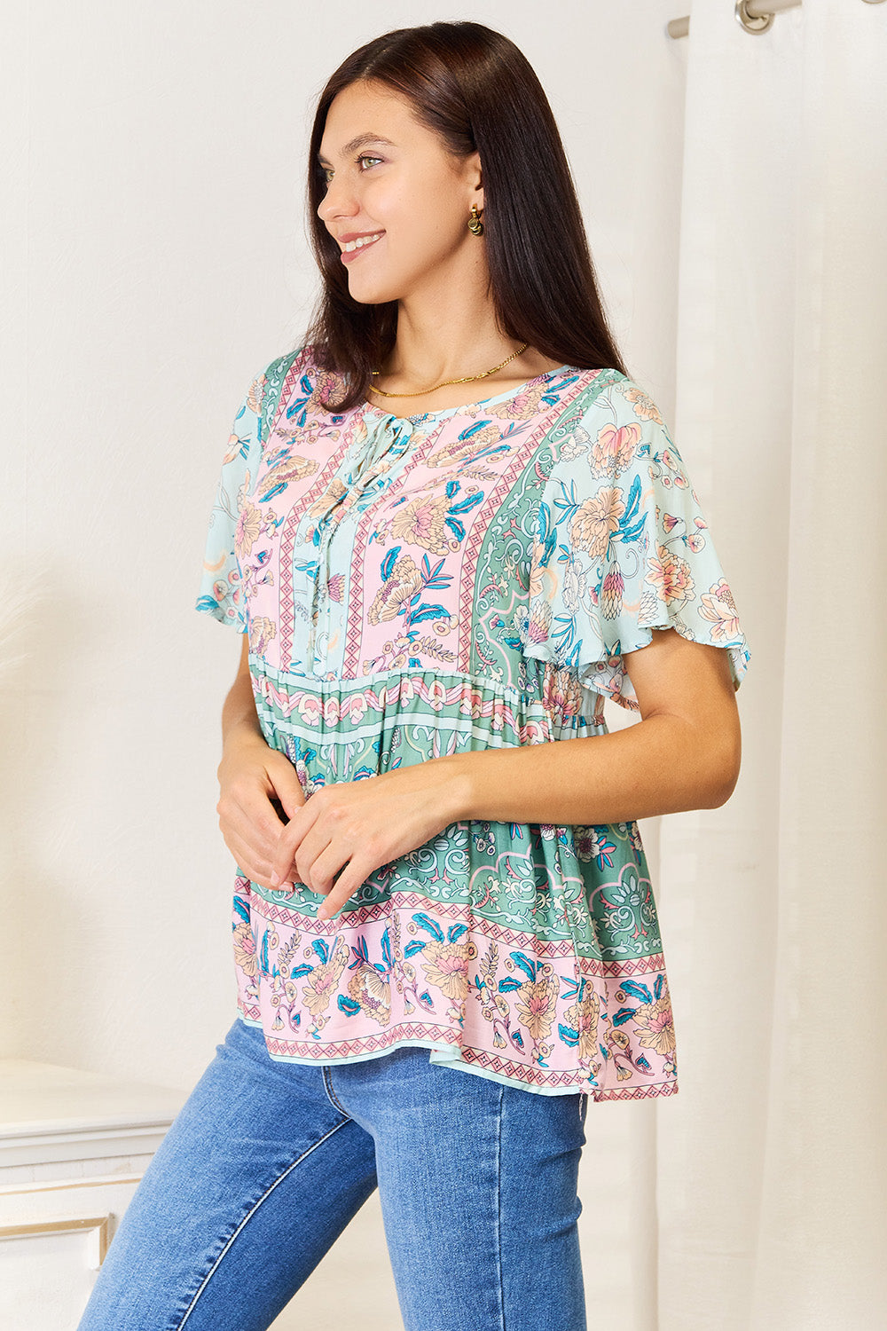 A person wearing a Perfee Floral Tie Neck Short Sleeve Blouse and denim jeans stands indoors, smiling slightly.