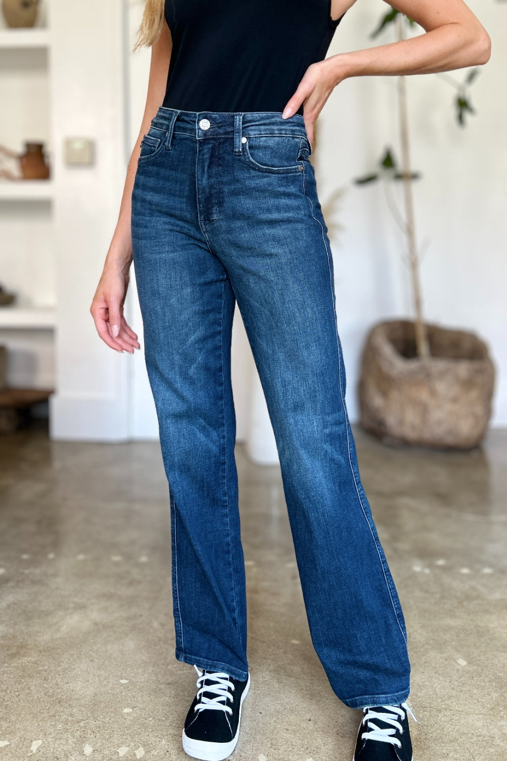 A person wearing a black top and Judy Blue Full Size Tummy Control Straight Jeans stands indoors with one hand on their hip. Their shoes are black with white soles. A plant and shelves are in the background.