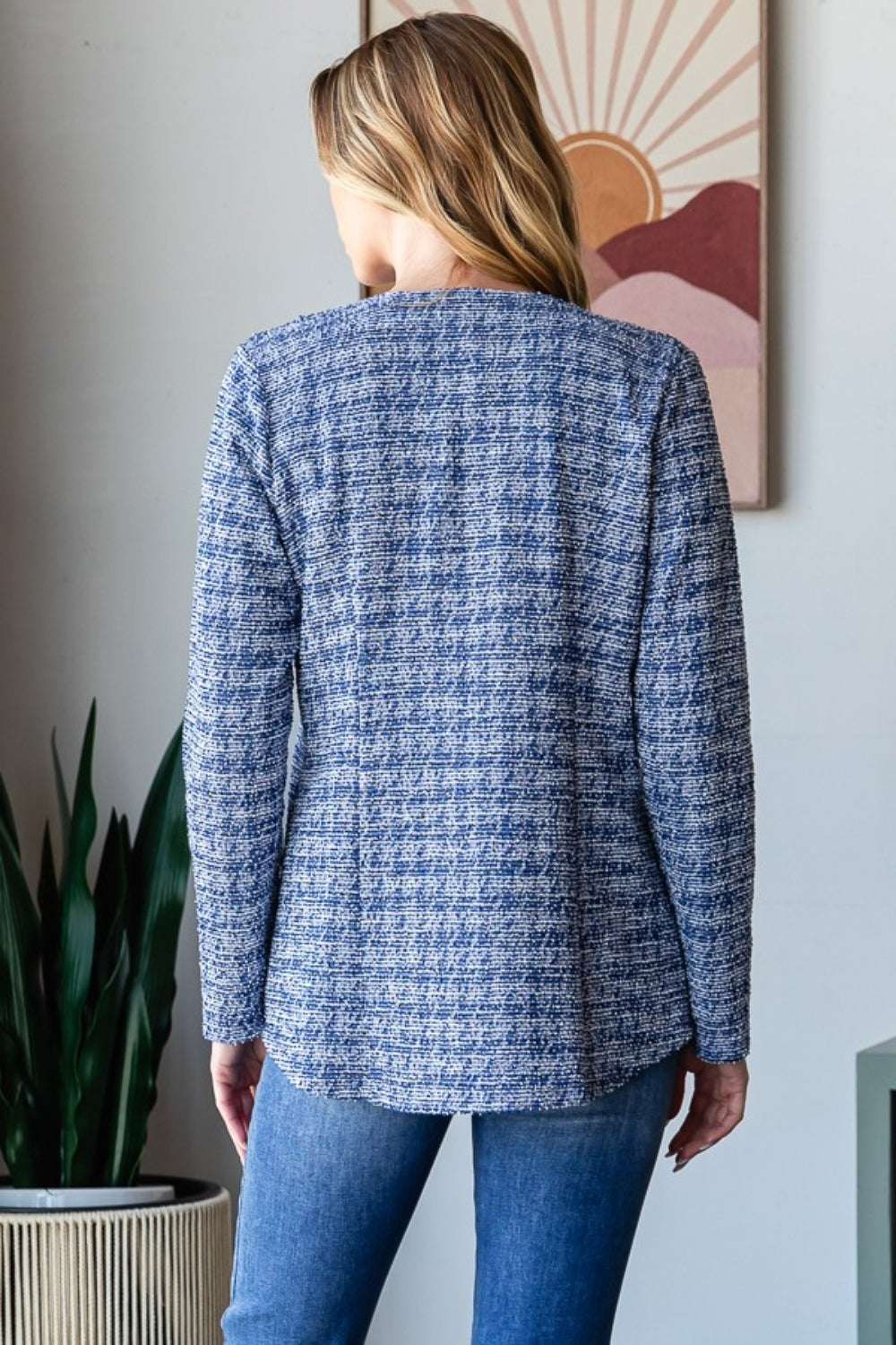 A woman with wavy blonde hair wears the Heimish Full Size Houndstooth Tweed Open Front Blazer in blue, paired with a white top and blue jeans. She stands in front of a wall adorned with framed art.