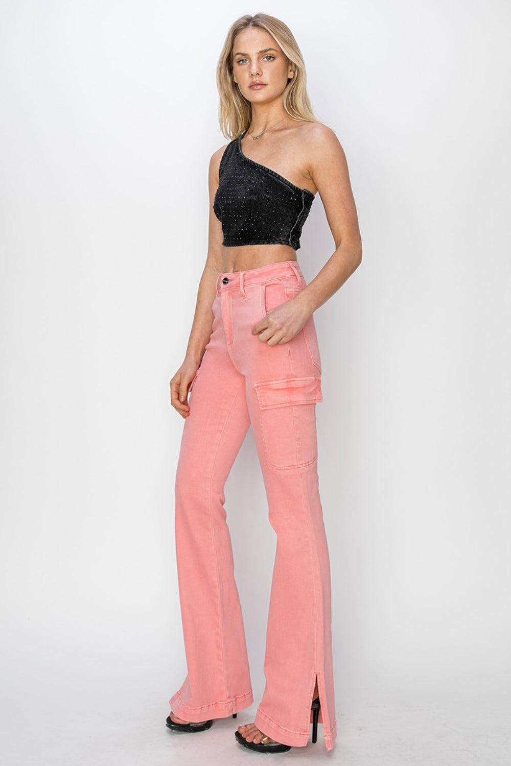 A person wearing RISEN Full Size High Rise Side Slit Cargo Bootcut Jeans, featuring a pink flared design with front pockets, stands against a plain background.