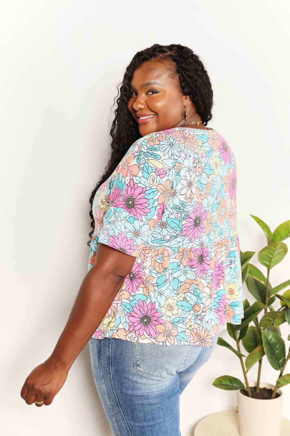 A person wearing a Double Take Floral Round Neck Babydoll Top and jeans stands with one hand in their pocket, with a plant in the background.