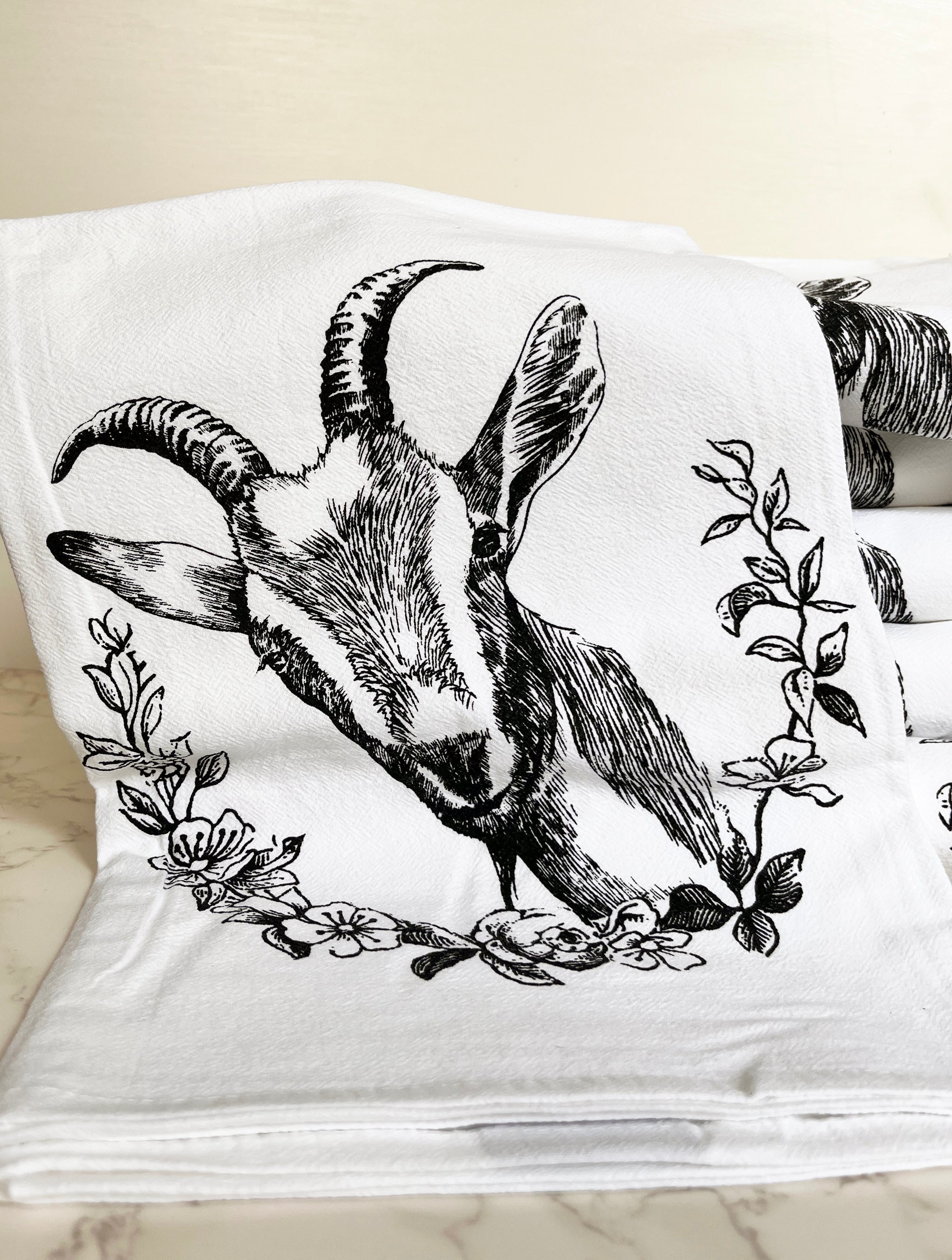 The Goat Cotton Kitchen Towel is a farmhouse-style cotton towel that is lint-free, showcasing a black goat illustration surrounded by floral designs on a crisp white background, beautifully presented on a marble surface.