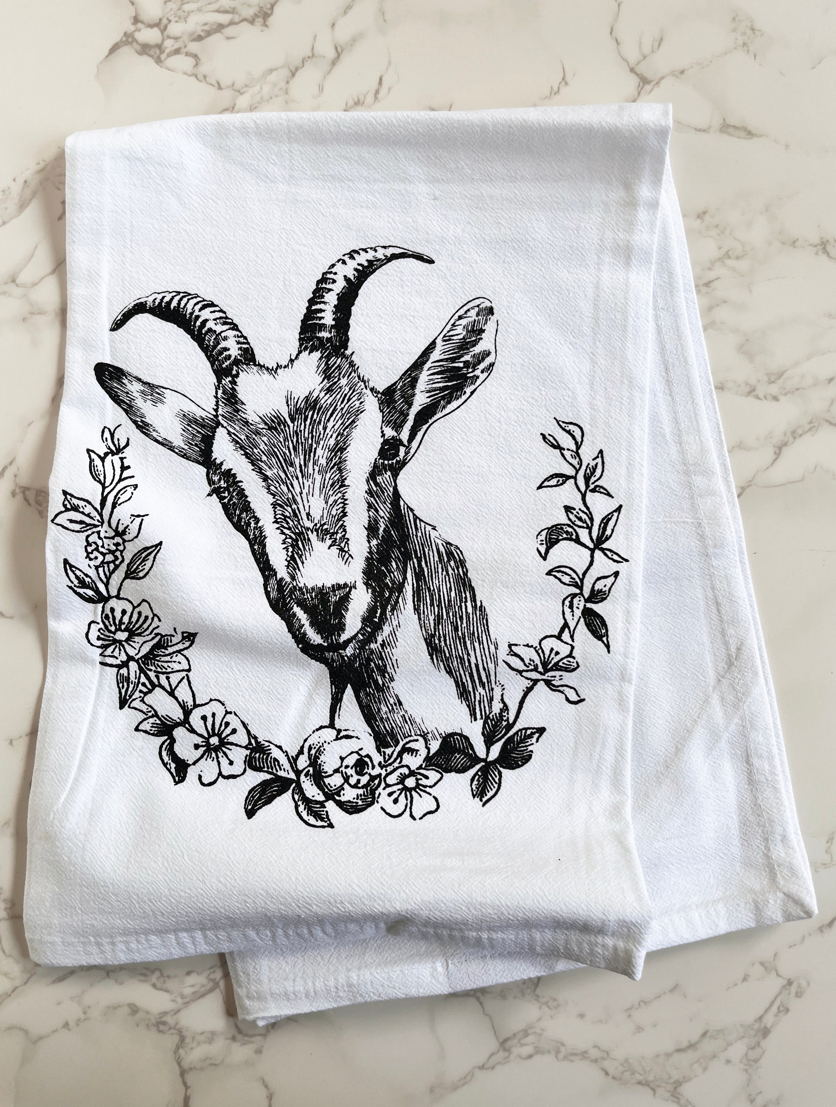 The Goat Cotton Kitchen Towel is a farmhouse-style cotton towel that is lint-free, showcasing a black goat illustration surrounded by floral designs on a crisp white background, beautifully presented on a marble surface.