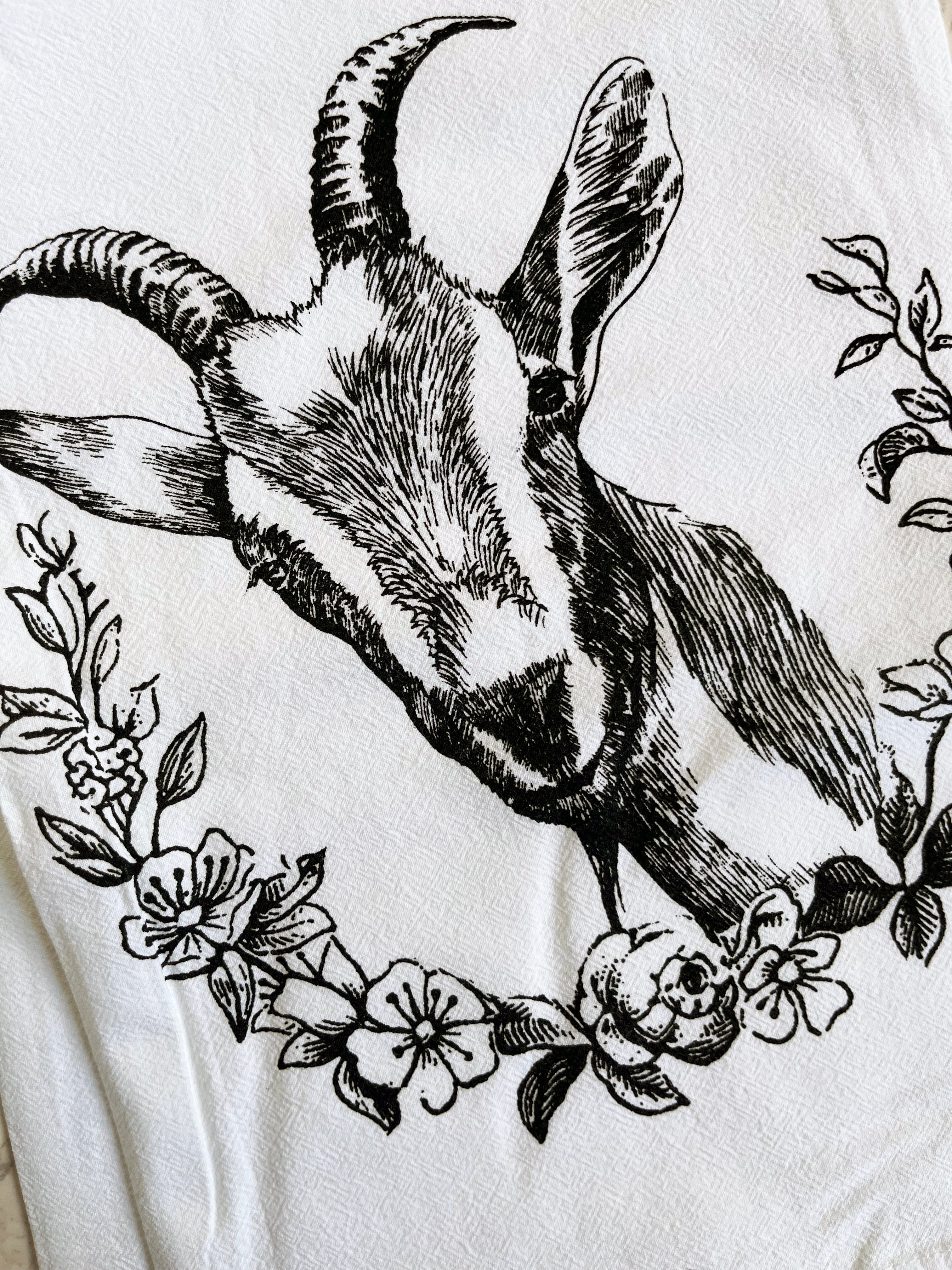 The Goat Cotton Kitchen Towel is a farmhouse-style cotton towel that is lint-free, showcasing a black goat illustration surrounded by floral designs on a crisp white background, beautifully presented on a marble surface.