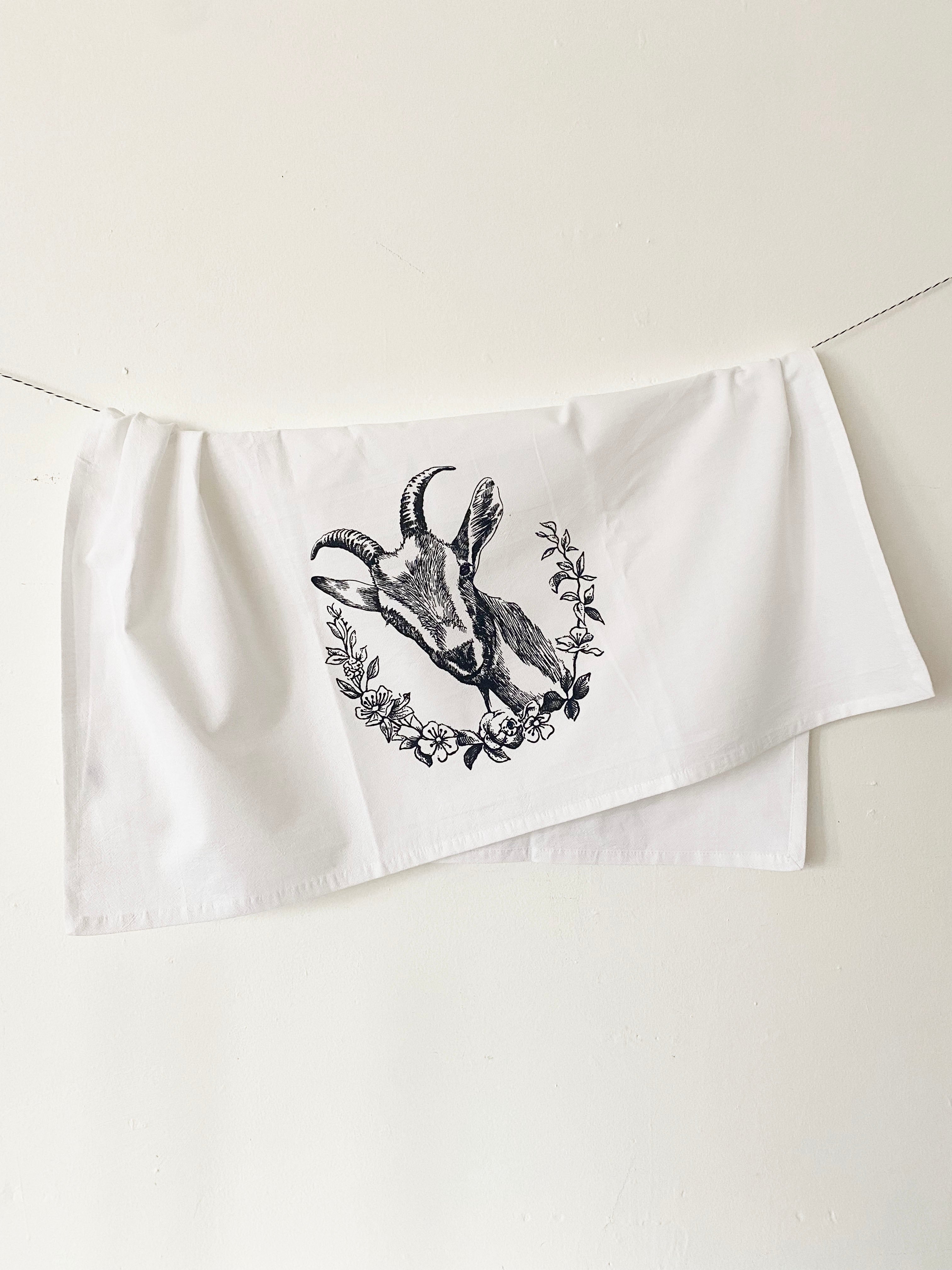 The Goat Cotton Kitchen Towel is a farmhouse-style cotton towel that is lint-free, showcasing a black goat illustration surrounded by floral designs on a crisp white background, beautifully presented on a marble surface.