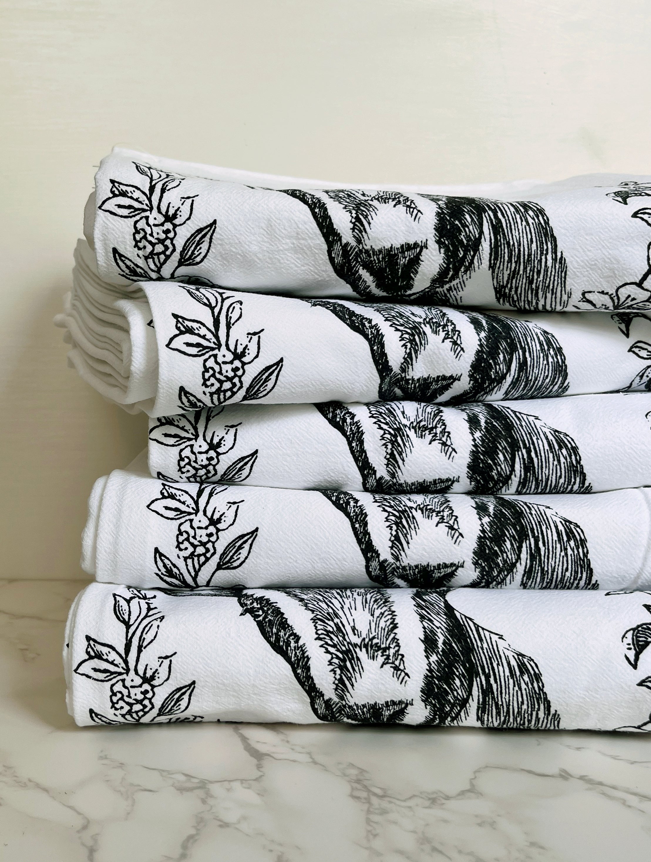 The Goat Cotton Kitchen Towel is a farmhouse-style cotton towel that is lint-free, showcasing a black goat illustration surrounded by floral designs on a crisp white background, beautifully presented on a marble surface.