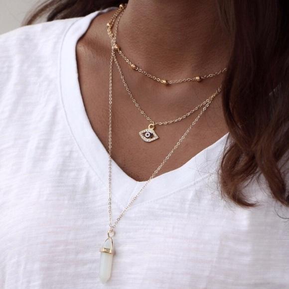 A person wearing the All Seeing Eye Multilayer Necklace with a gold eye-shaped pendant and another featuring a vertically elongated natural stone, possibly opal or amethyst, layered over a crisp white shirt.