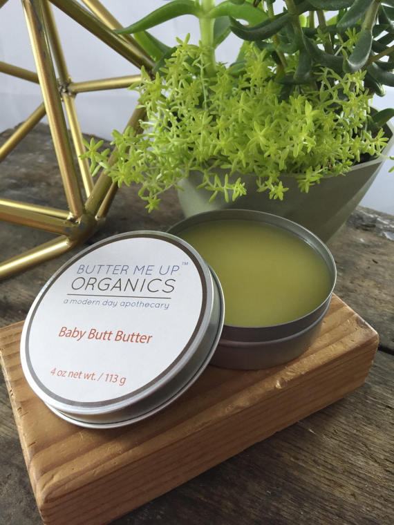 A tin labeled "Baby Butt Butter- Organic Diaper Cream," made from natural ingredients to soothe diaper rash, sits elegantly on a stone slab with wooden sticks on the side.