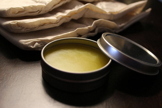 A tin labeled "Baby Butt Butter- Organic Diaper Cream," made from natural ingredients to soothe diaper rash, sits elegantly on a stone slab with wooden sticks on the side.