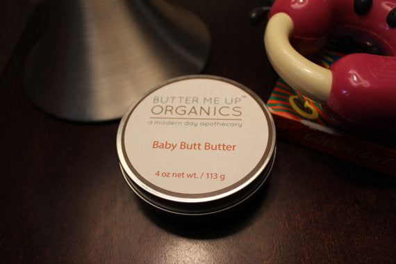 A tin labeled "Baby Butt Butter- Organic Diaper Cream," made from natural ingredients to soothe diaper rash, sits elegantly on a stone slab with wooden sticks on the side.