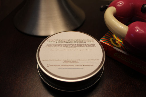 A tin labeled "Baby Butt Butter- Organic Diaper Cream," made from natural ingredients to soothe diaper rash, sits elegantly on a stone slab with wooden sticks on the side.