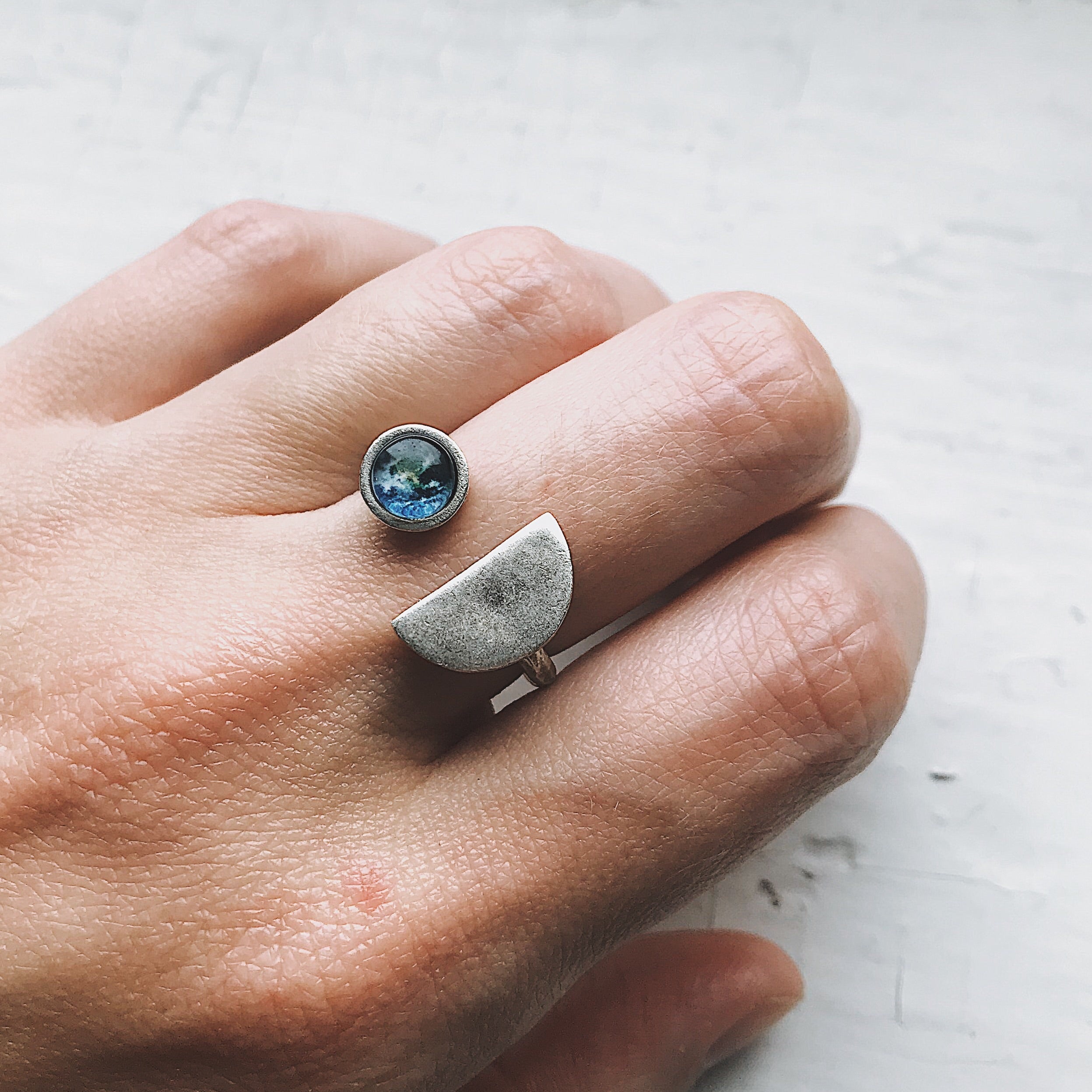 A hand displays two exquisite handcrafted jewelry pieces: one, the Earth Rise Ring, has a small Earth image reminiscent of Apollo 8, and the other showcases a simple, elegant half-circle shape.