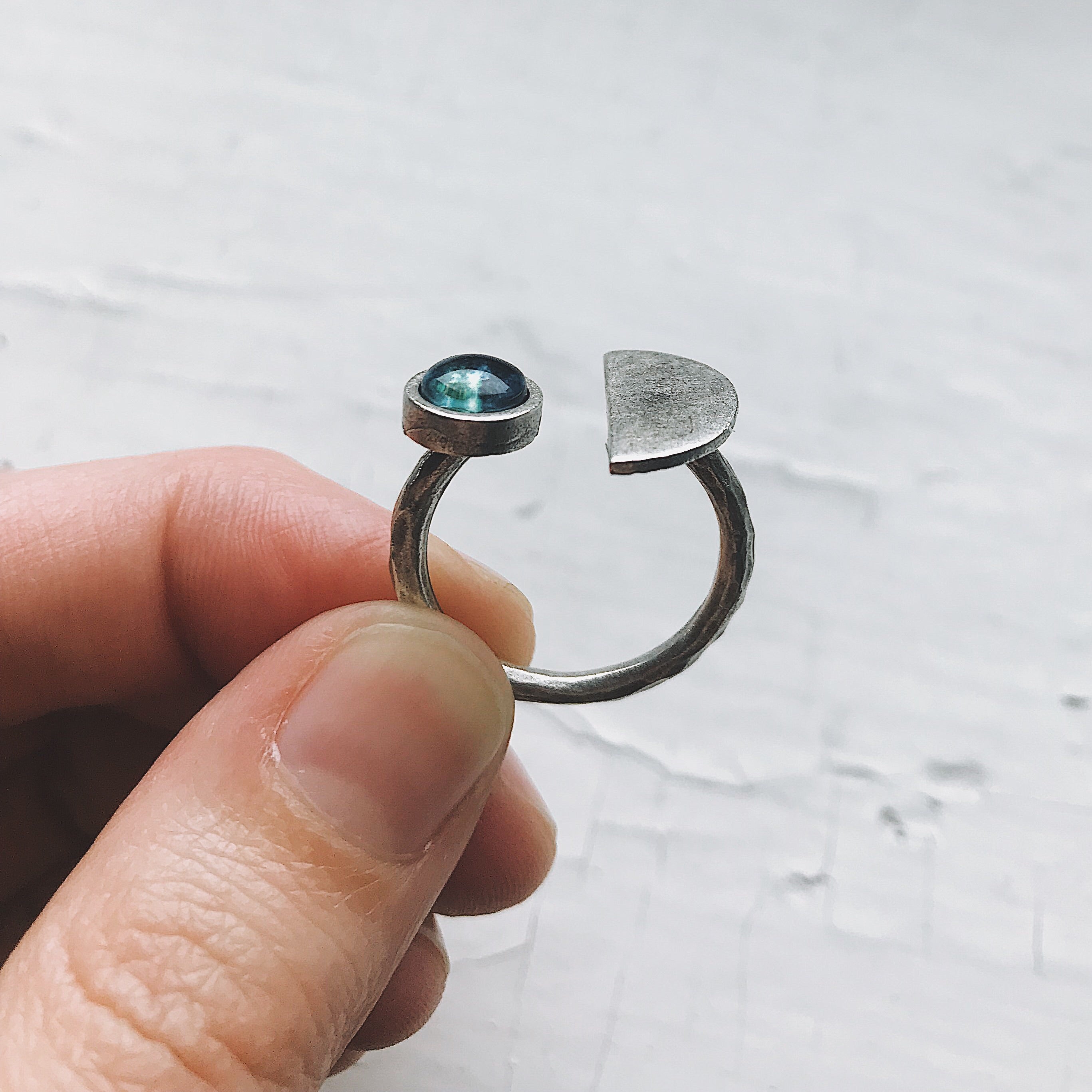 The Earth Rise Ring, a rhodium-plated piece featuring a blue stone in a half-moon shape, rests on textured wood, its handcrafted elegance highlighted against a blurred green background.