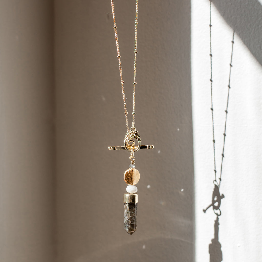 The Labradorite Lariat Necklace is a stunning gold necklace featuring a toggle clasp, a vertical rectangular labradorite stone pendant, and a round bead above it.