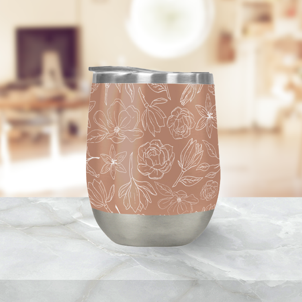 The Copper Magnolia Wine Tumbler, featuring a stainless steel build and floral design, sits on a marble surface against a blurred indoor background, highlighting its elegant style and efficient vacuum insulation.