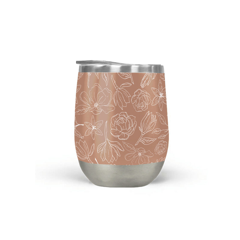 The Copper Magnolia Wine Tumbler, featuring a stainless steel build and floral design, sits on a marble surface against a blurred indoor background, highlighting its elegant style and efficient vacuum insulation.