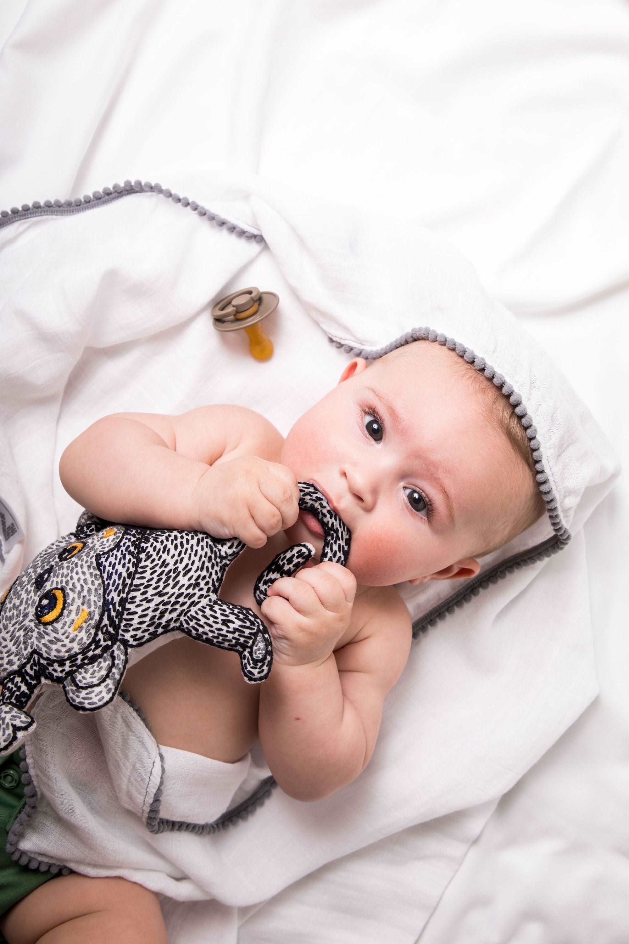 A white 3 Pc Newborn Essential Set - Hooded Towel, Swaddle + Toy Rattle by Ivory Meleager on top of a bed.