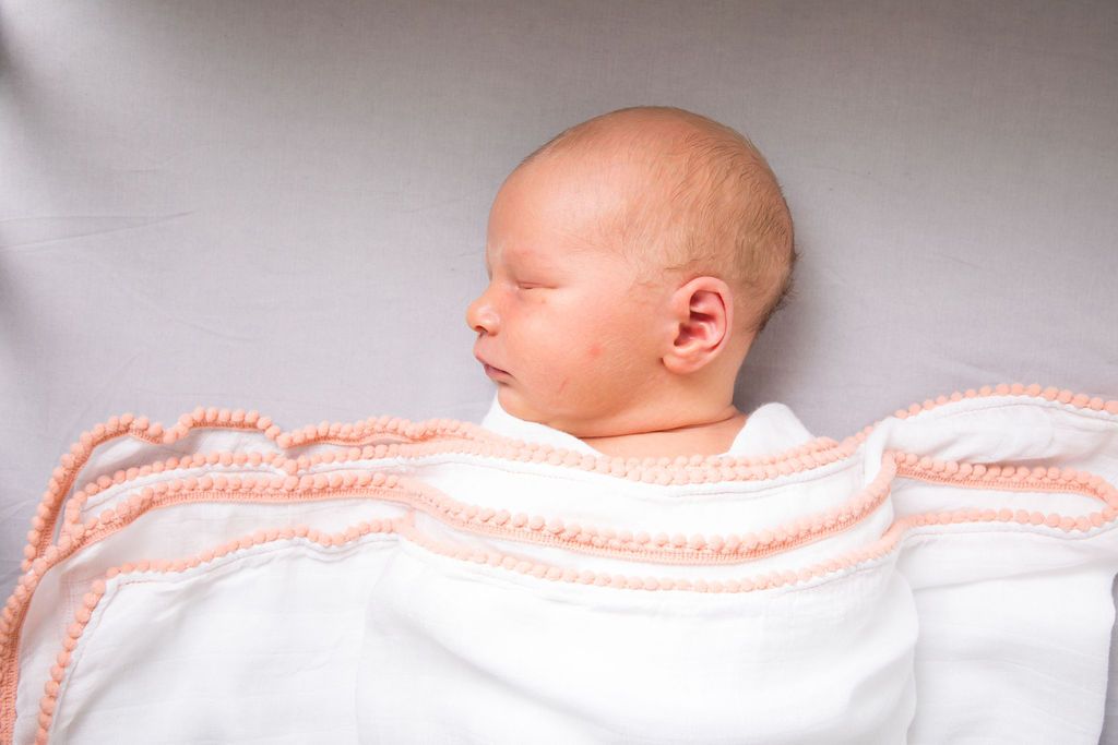 A white 3 Pc Newborn Essential Set - Hooded Towel, Swaddle + Toy Rattle by Ivory Meleager on top of a bed.