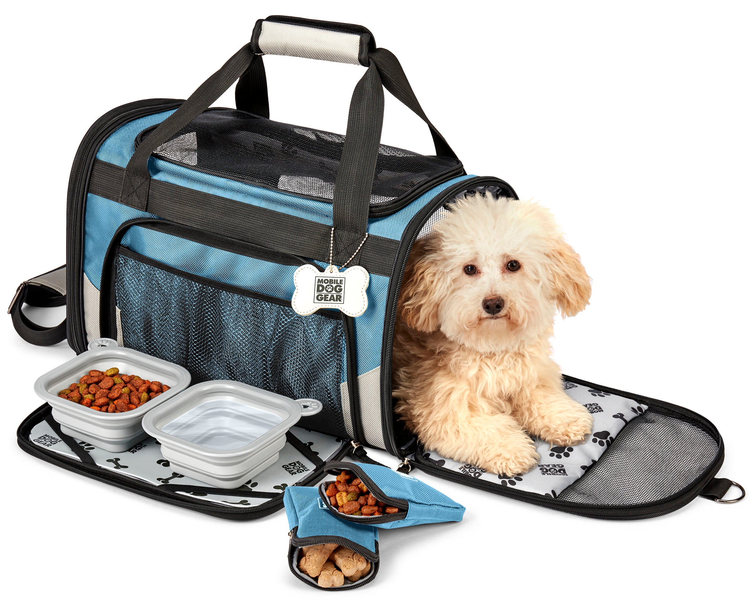 A small dog sits comfortably in a Mobile Dog Gear Pet Carrier Plus, complete with food and water bowls nearby. A speech bubble above says, "This is MY carrier!