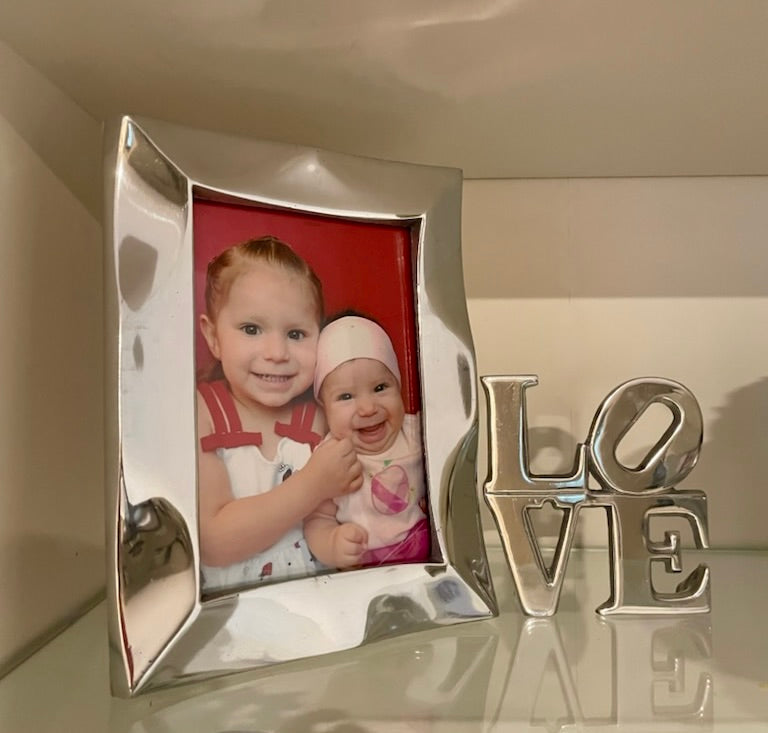 The Stylish Picture Frame is a minimalist design crafted from recycled aluminum, showcasing a silver finish with a plain brown backing and supported by a wooden stand at the back.