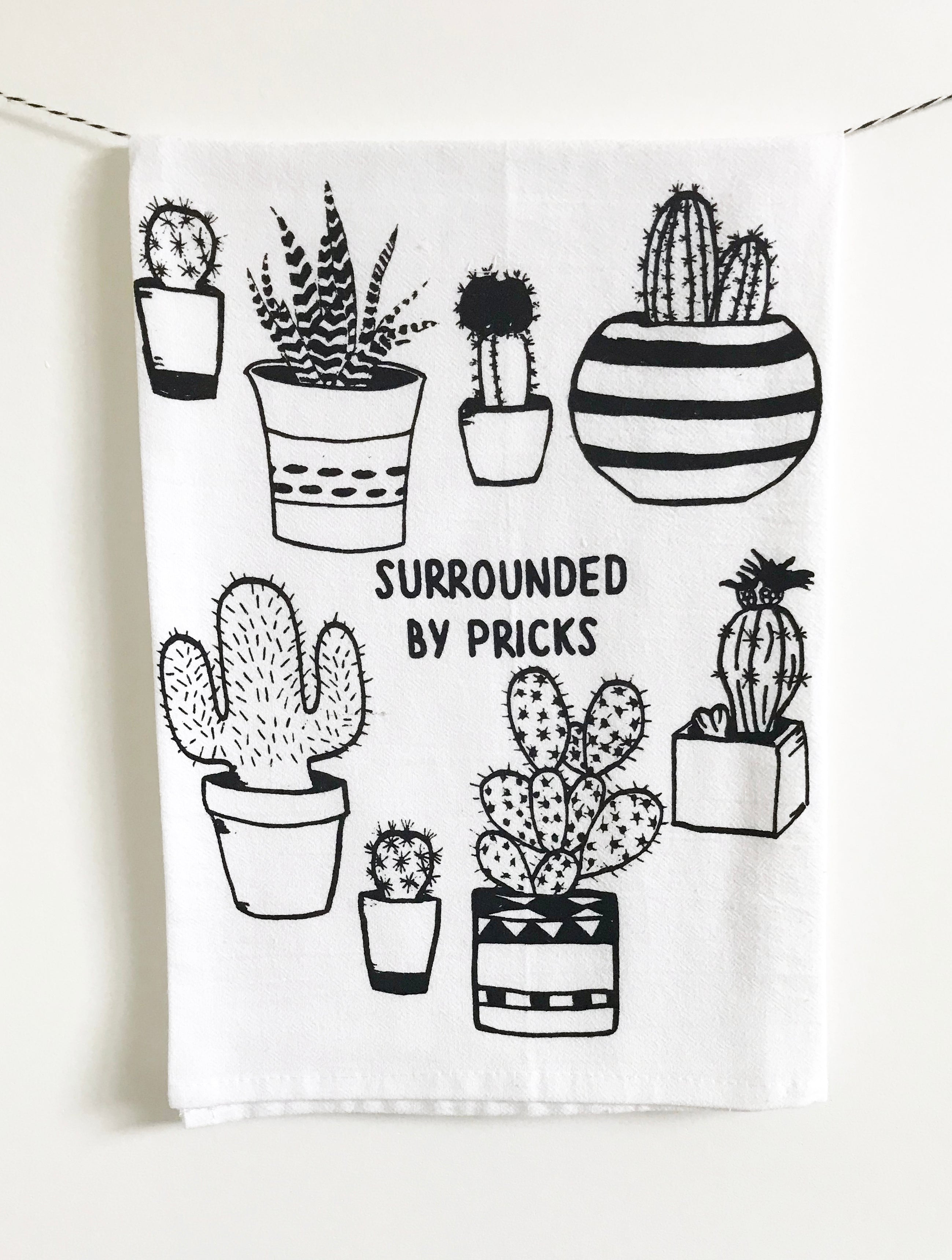 Introducing the "Surrounded by Pricks Cotton Kitchen Towel," a lint-free cotton towel featuring black line drawings of various potted cacti accompanied by the playful text "Surrounded by Pricks." Ideal for an eco-friendly kitchen, this towel makes a wonderful gift for plant enthusiasts who value humor and sustainability.