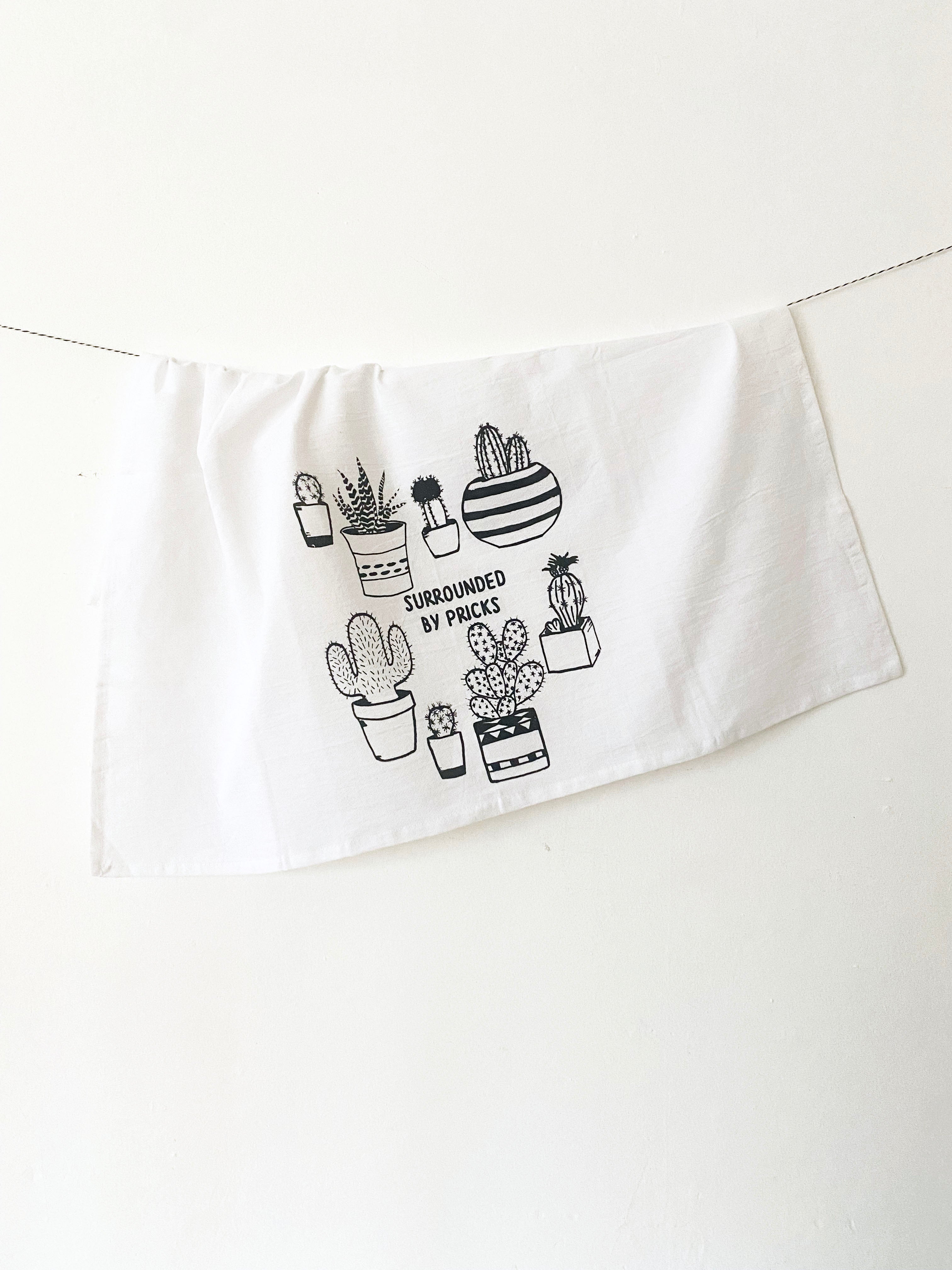 Introducing the "Surrounded by Pricks Cotton Kitchen Towel," a lint-free cotton towel featuring black line drawings of various potted cacti accompanied by the playful text "Surrounded by Pricks." Ideal for an eco-friendly kitchen, this towel makes a wonderful gift for plant enthusiasts who value humor and sustainability.