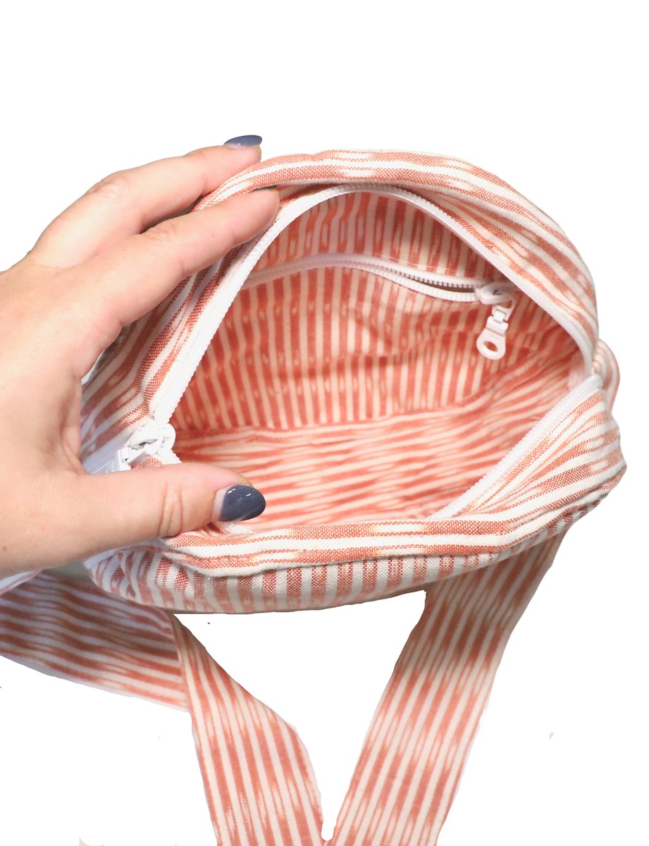 Person opening a round crossbody bag with a blue pattern, while wearing an orange dress adorned with white floral designs, reminiscent of fair trade craftsmanship.