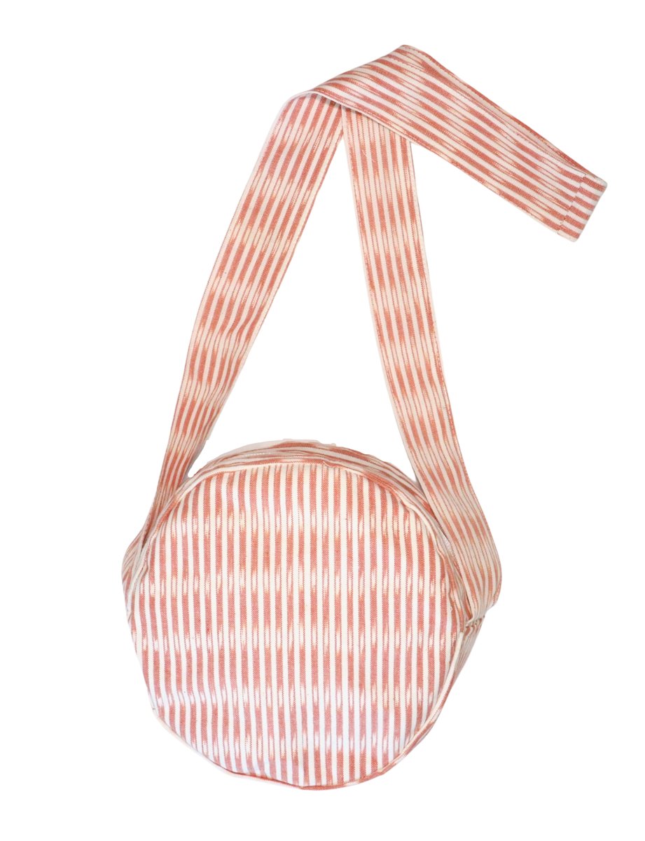 Dressed in a striped outfit, an individual carefully opens a small Round Crossbody Bag from the Multiple Prints Available collection. This charming and handwoven cotton bag with a shoulder strap enhances their look with style and practicality, showcasing their dedication to fair trade fashion.