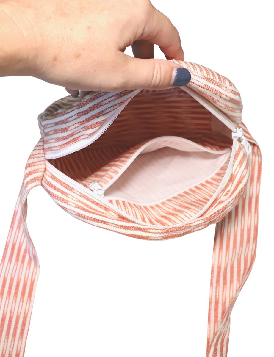 Person opening a round crossbody bag with a blue pattern, while wearing an orange dress adorned with white floral designs, reminiscent of fair trade craftsmanship.