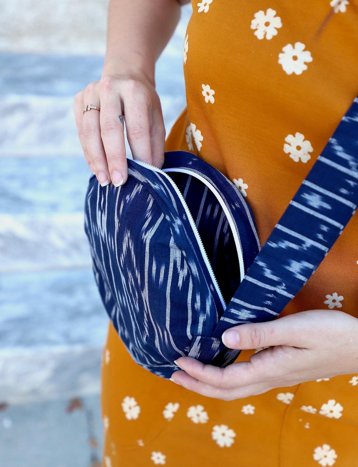 Person opening a round crossbody bag with a blue pattern, while wearing an orange dress adorned with white floral designs, reminiscent of fair trade craftsmanship.