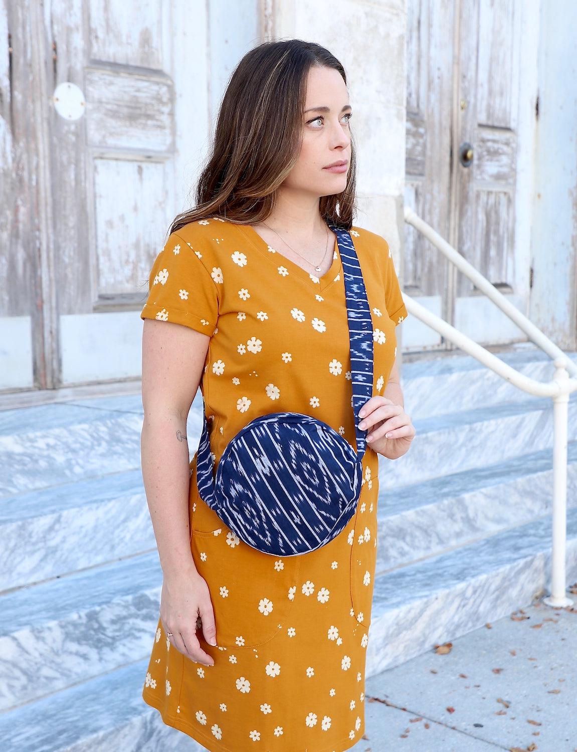 Person opening a round crossbody bag with a blue pattern, while wearing an orange dress adorned with white floral designs, reminiscent of fair trade craftsmanship.