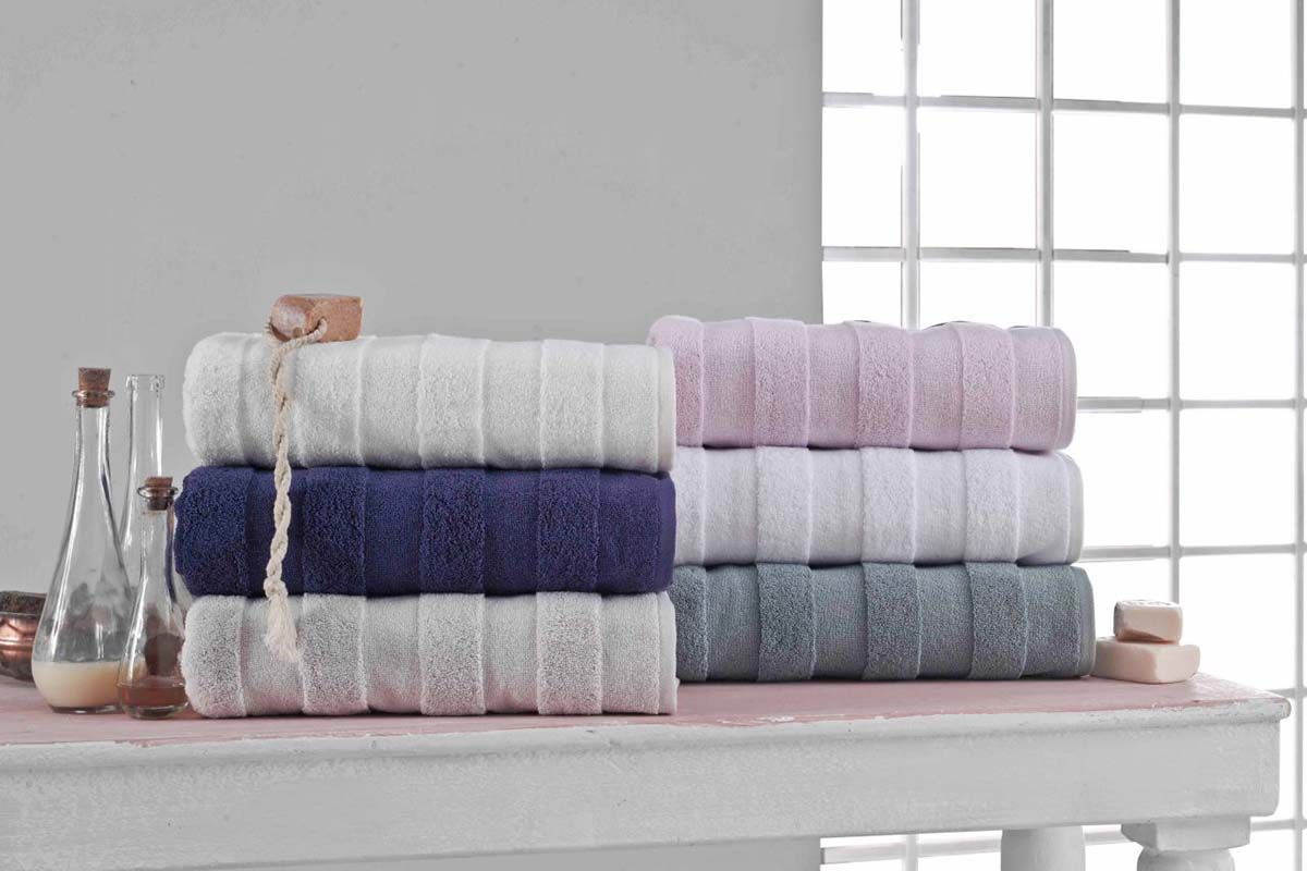 The Apogee Collection 3 PK Towels Set, featuring luxurious towels in white, blue, pink, and gray, is expertly arranged on a shelf. Nearby, small bottles and bars of soap complement the display with their decorative touch. Crafted from ultra-soft cotton, these towels enhance any bathroom experience with their plush texture.