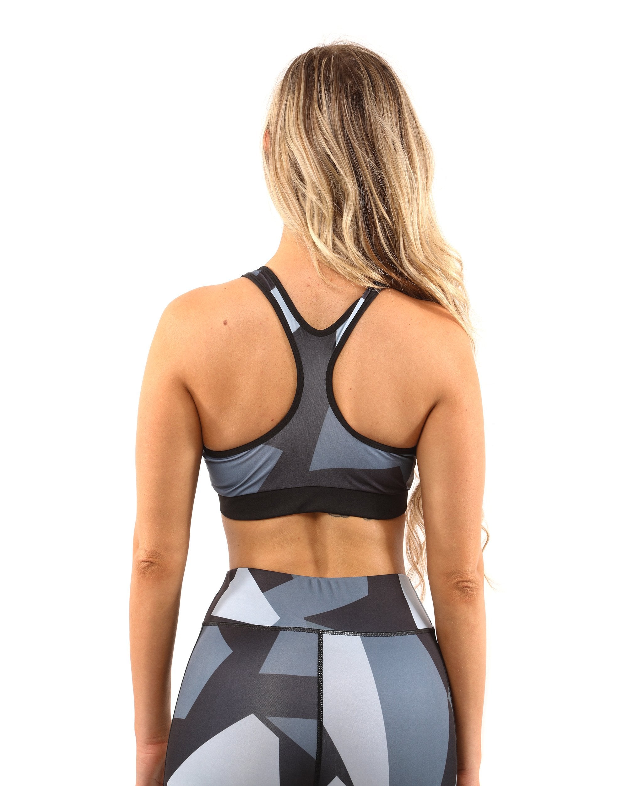 A person with long blonde hair is wearing the Bondi Sports Bra - Black/Grey, featuring a geometric-patterned compression fit and racerback design, accompanied by matching leggings, standing against a white background.