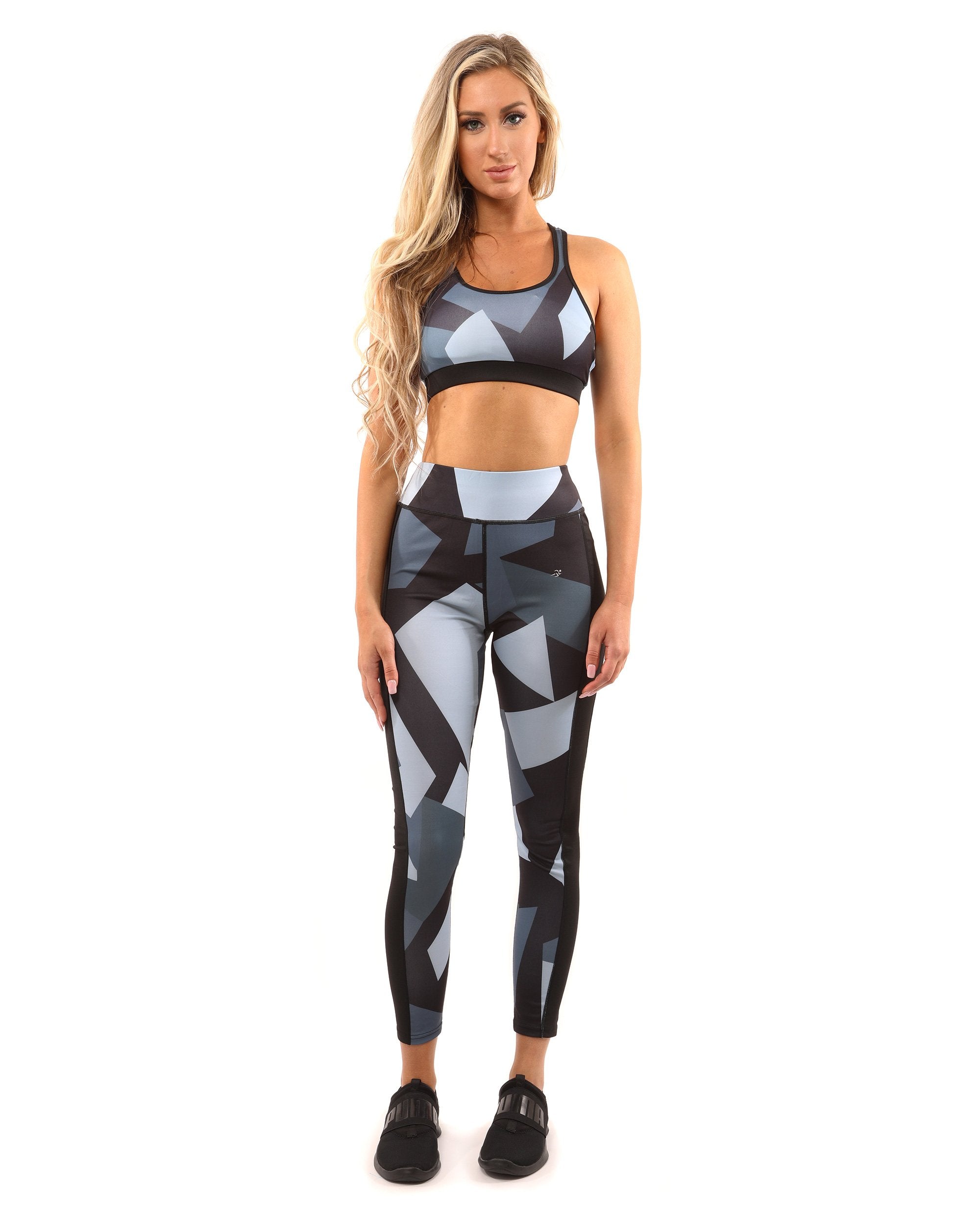 A person with long blonde hair is wearing the Bondi Sports Bra - Black/Grey, featuring a geometric-patterned compression fit and racerback design, accompanied by matching leggings, standing against a white background.