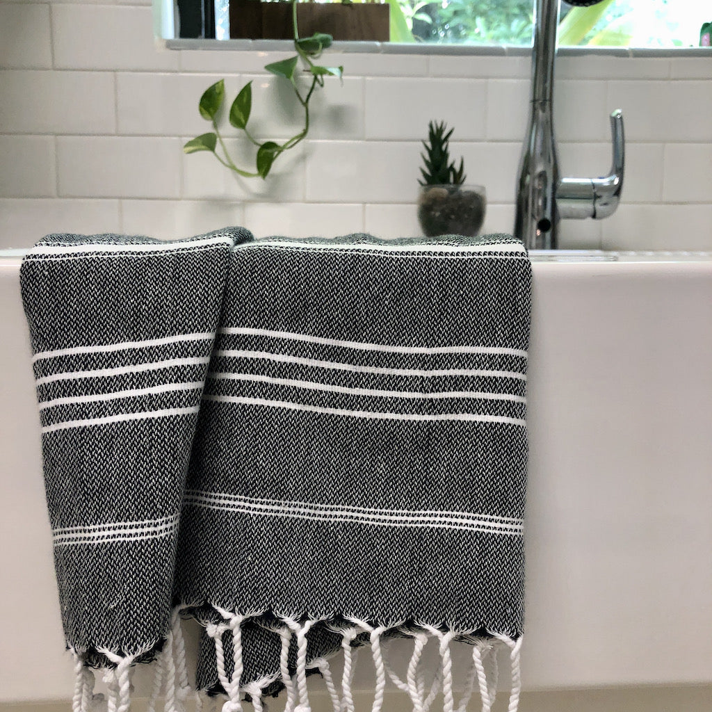 The Classic Turkish Hand Towels in five elegant striped designs, featuring shades of blue, brown, beige, navy, and grey, are beautifully displayed. Made from 100% Turkish cotton and adorned with white fringe on one end, they offer exceptional absorbency for all your needs.