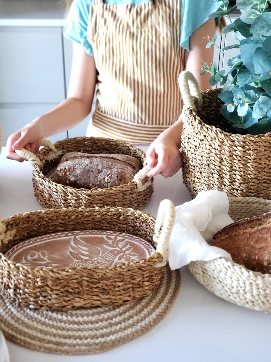 The Bird Oval Bread Warmer & Basket is an expertly crafted product, featuring an oval terracotta platter adorned with a white bird design, set within a handcrafted basket with two loop handles by Fair Trade artisans.