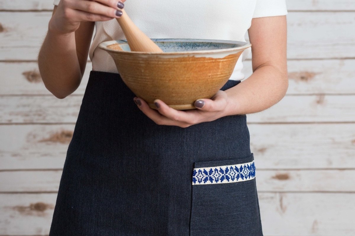 A person stands in a kitchen, hands on hips, wearing the Farah Half Apron - Blue over a white shirt and dark skirt with a blue patterned pocket.