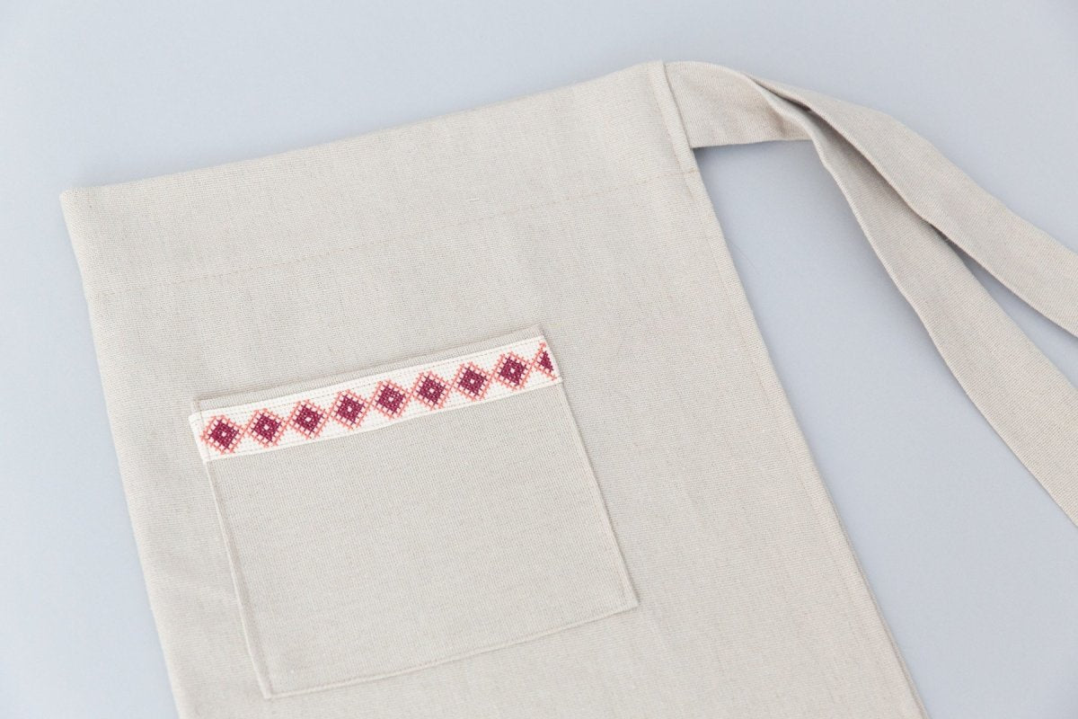 A person wearing a cream Farah Half Apron with a wooden spoon tucked into the front pocket. This apron showcases an empowering red and white embroidered tatreez motif.