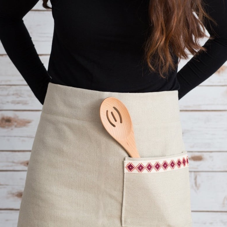 A person wearing a cream Farah Half Apron with a wooden spoon tucked into the front pocket. This apron showcases an empowering red and white embroidered tatreez motif.