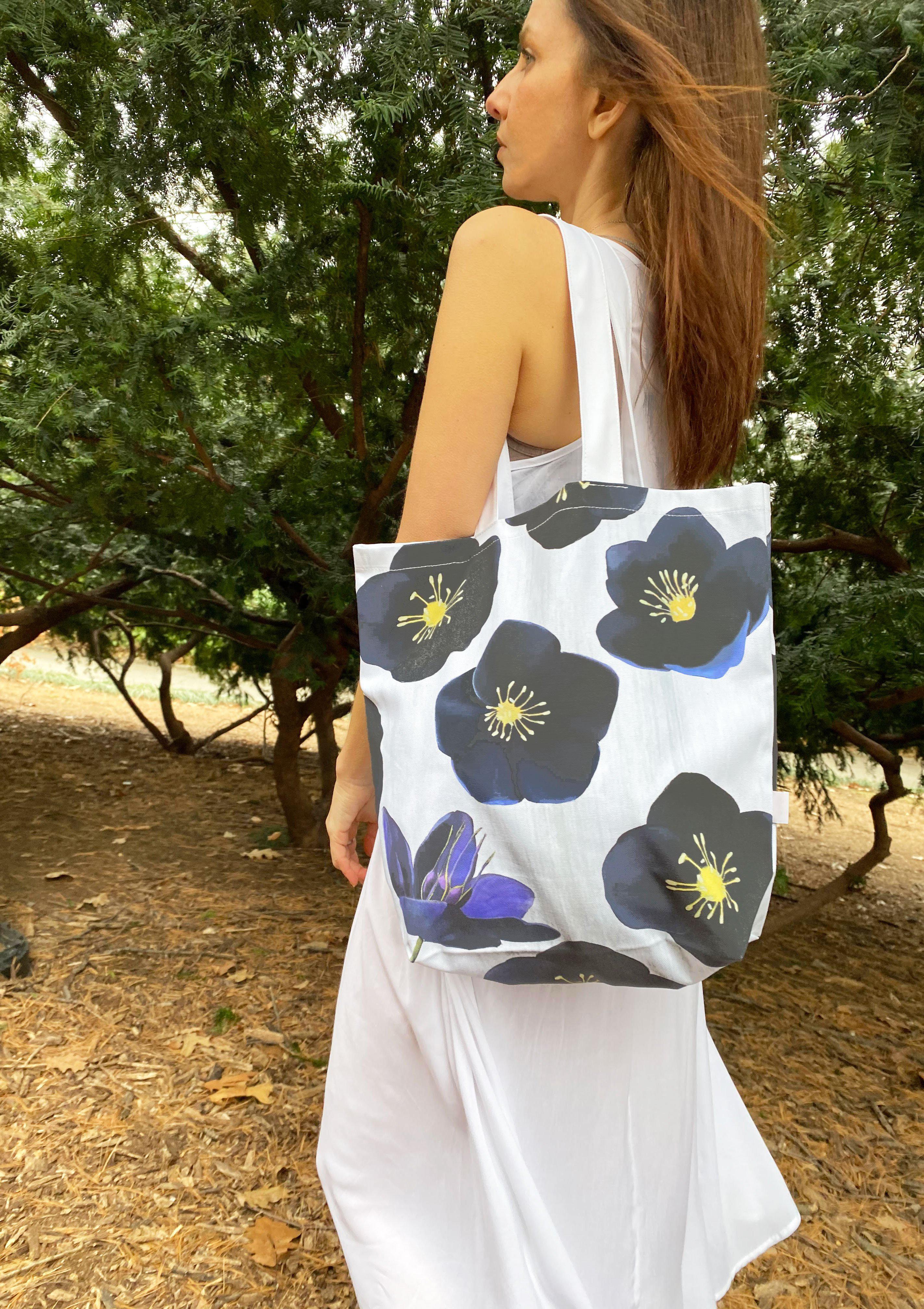 The Tote Bag: Black Hellebore on Snow, a chic cotton canvas piece with an elegant floral design, hangs on a doorknob beside a bouquet of Black Hellebore flowers. An umbrella featuring the same floral pattern leans gracefully against the wall.