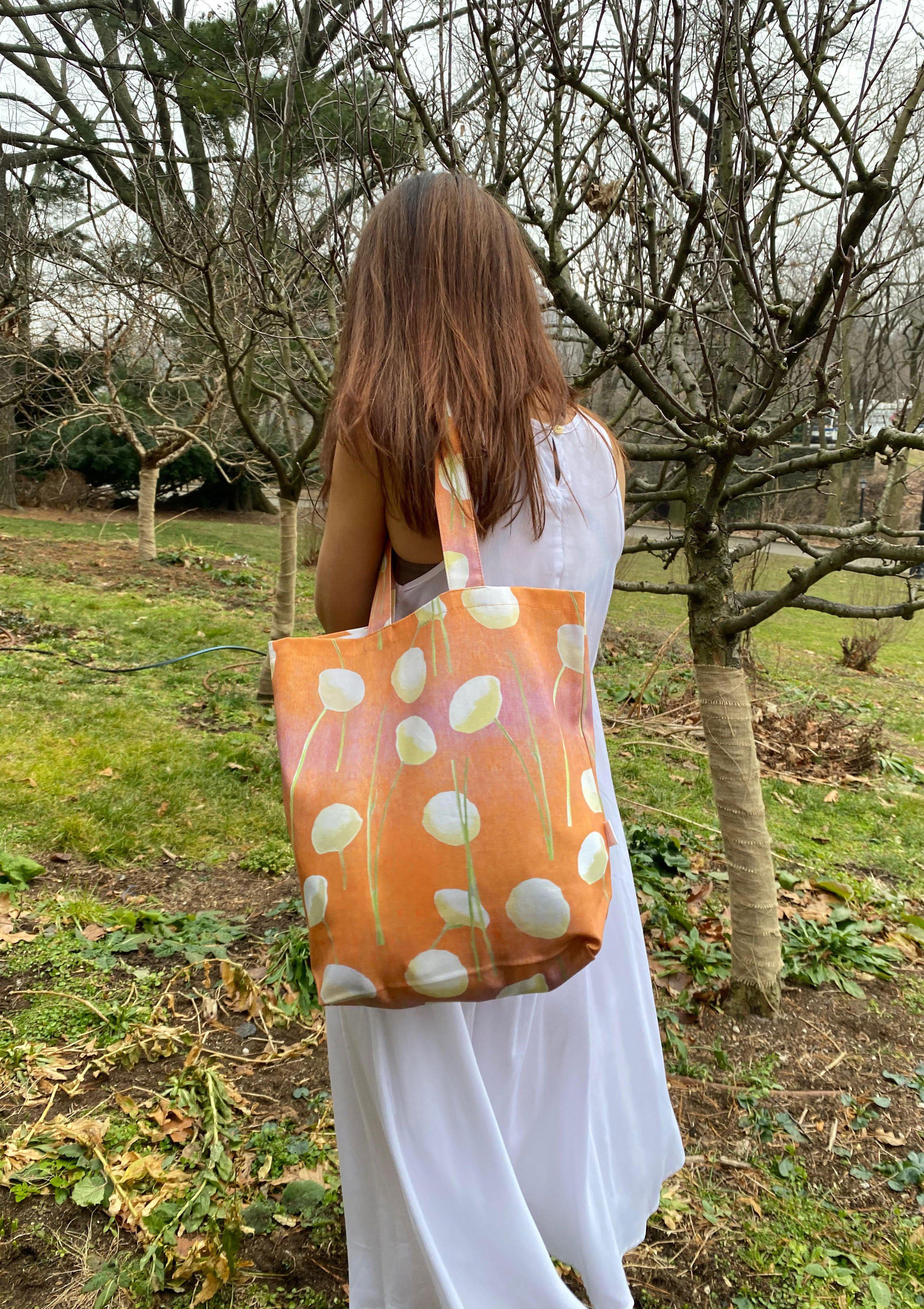 The "Tote Bag: Cream Flowers on Orange," made from stylish cotton canvas and featuring floral patterns, hangs on a doorknob beside a folded umbrella. An oil-painted chrysanthemum bouquet rests against the closed door.