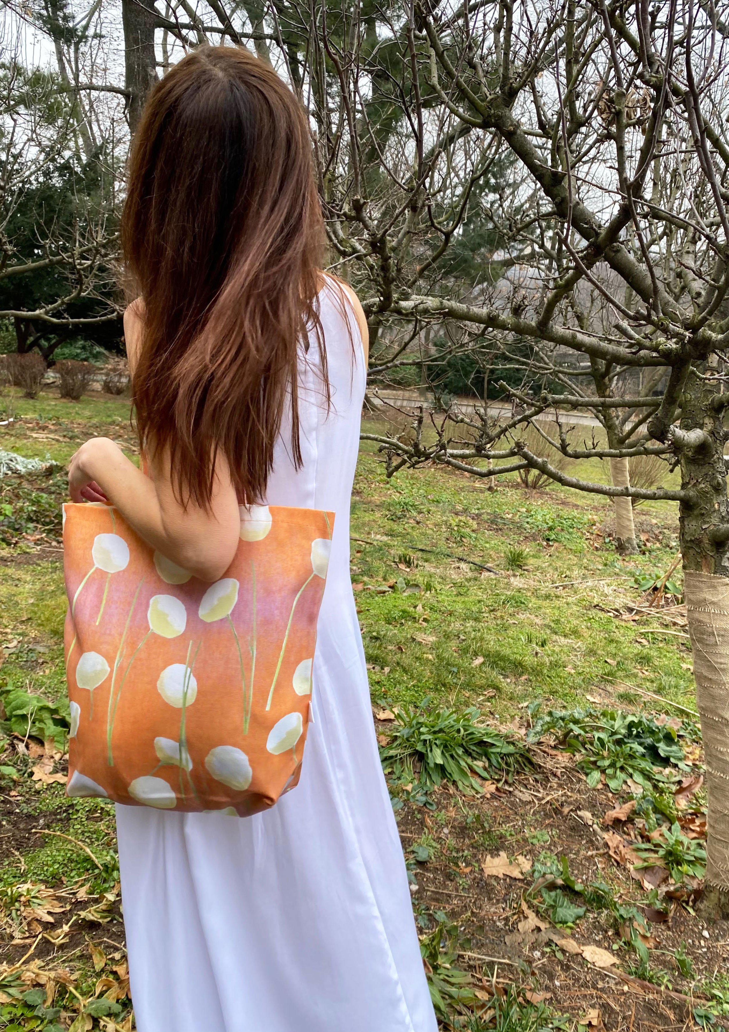 The "Tote Bag: Cream Flowers on Orange," made from stylish cotton canvas and featuring floral patterns, hangs on a doorknob beside a folded umbrella. An oil-painted chrysanthemum bouquet rests against the closed door.